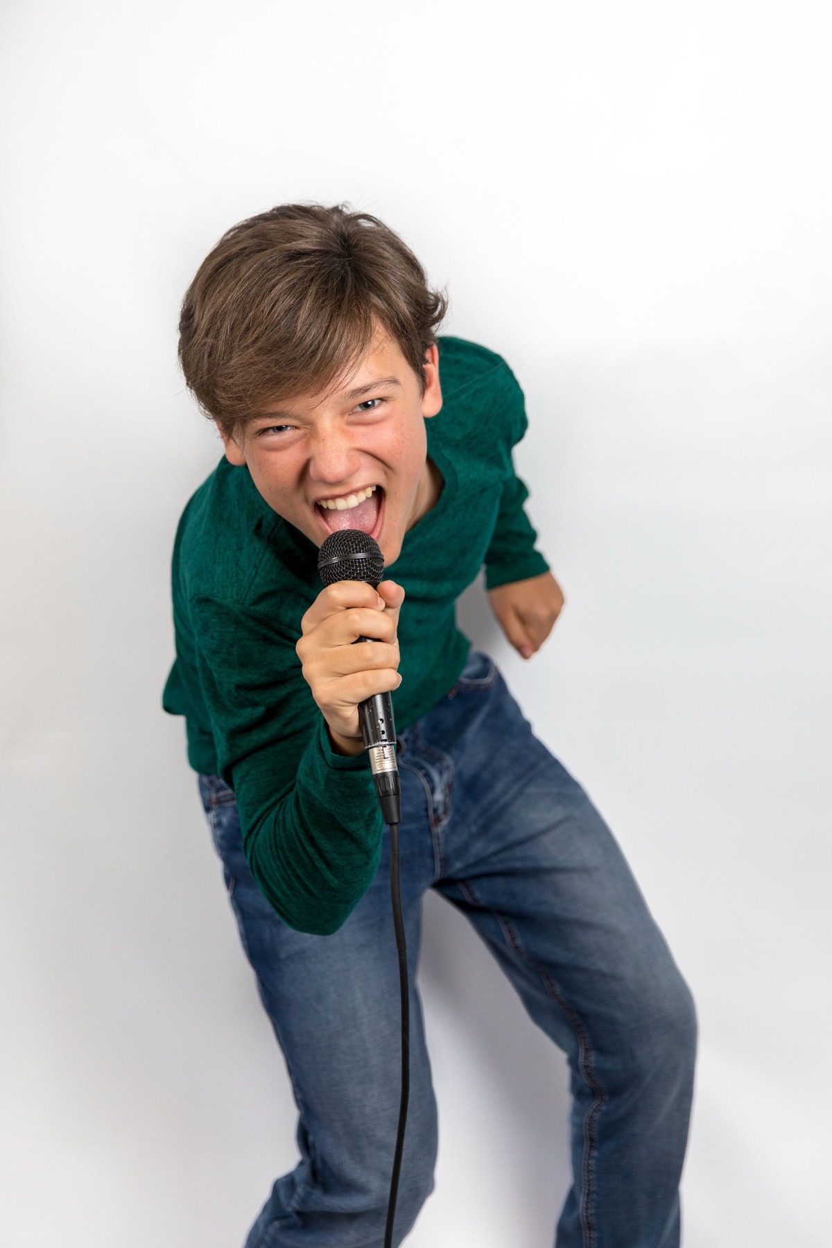 Person singing into a microphone, leaning forward in a green shirt and jeans against a white background.
